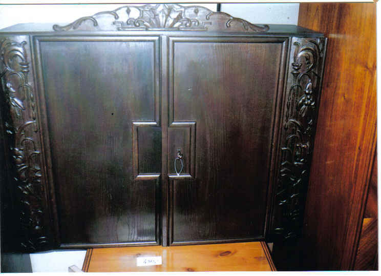 Ciemnobrązowa, dwudrzwiowa szafa z rzeźbionymi zdobieniami na krawędziach, stojąca na drewnianym blacie.